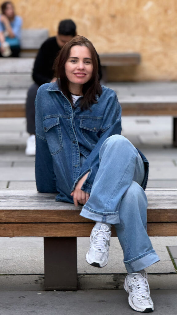 "A girl wearing a denim jacket and jeans sits comfortably on a wooden bench, smiling at the camera with a relaxed expression."